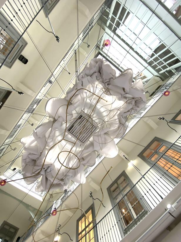 Blick von unten auf eine Kunstinstallation aus weißen Stoffbahnen und goldenen Drähten in einem Gebäude.
