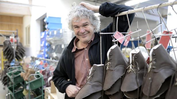 Ein lächelnder Mann steht in einer Werkstatt neben einer Kleiderstange mit Schuhen.