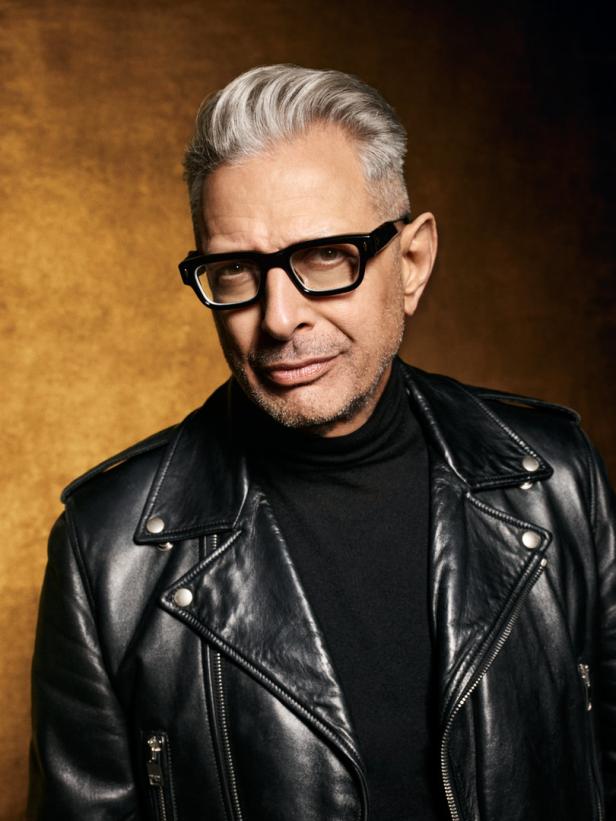Porträt von Jeff Goldblum mit Brille und Lederjacke vor goldenem Hintergrund.