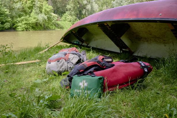 Ein umgedrehtes Kanu liegt am Ufer, daneben Rucksäcke und ein Erste-Hilfe-Kasten.