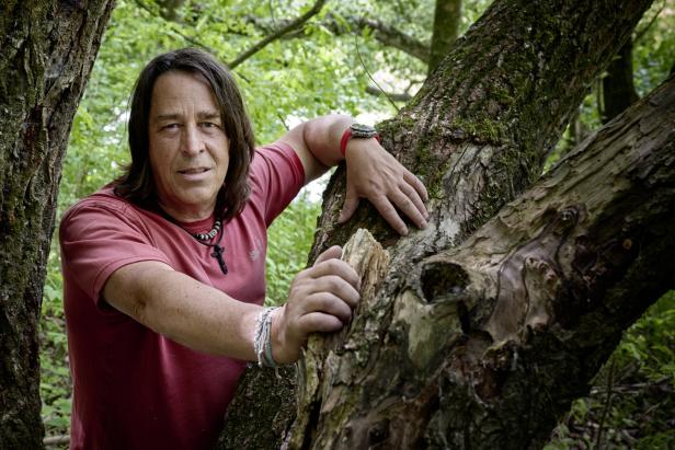 Ein Mann mit langem Haar lehnt an einem Baum im Wald.