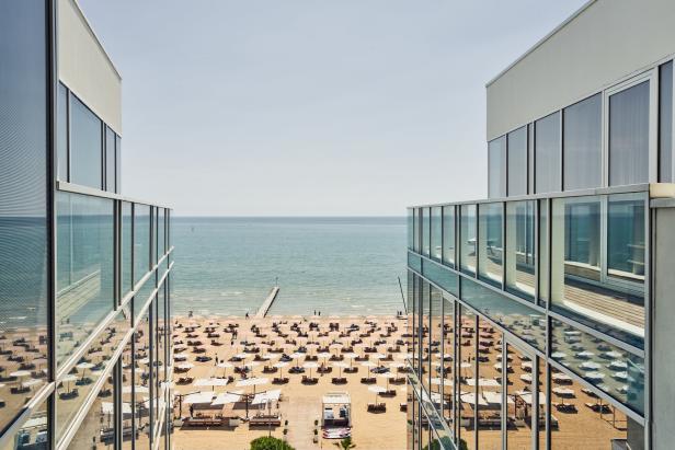 Blick zwischen zwei Gebäuden auf einen Strand mit vielen Sonnenschirmen und das Meer.