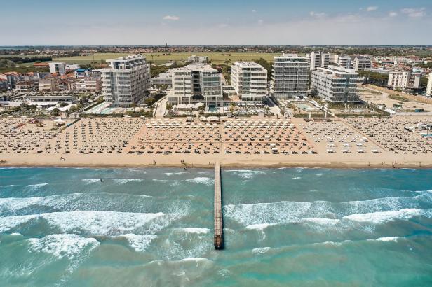 Luftaufnahme eines Strandes mit Liegestühlen, einem Pier und modernen Hotelgebäuden im Hintergrund.