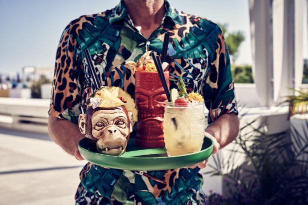 Ein Mann im bunten Hemd hält ein Tablett mit drei exotischen Cocktails.