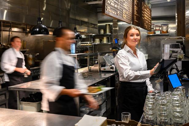 Eine Frau bedient eine Kasse in einer Restaurantküche, während zwei Köche im Hintergrund arbeiten.