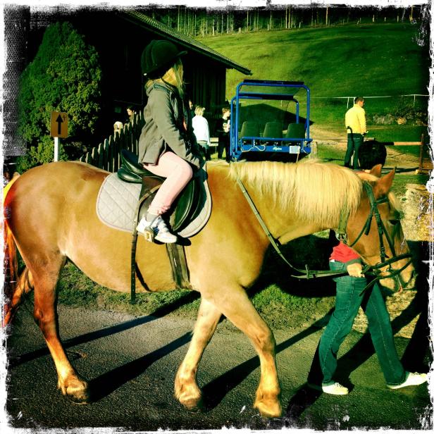 Ein Mädchen mit Reithelm sitzt auf einem hellbraunen Pferd, das von einer Person geführt wird.