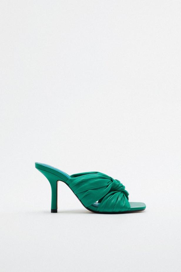 Ein grüner, hochhackiger Mules-Schuh mit Knotendetail steht vor einem weißen Hintergrund.