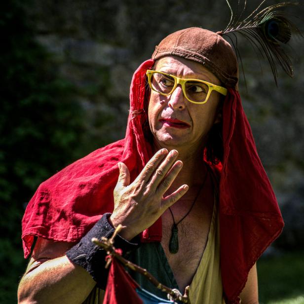 Ein Mann mit gelber Brille, rotem Lippenstift und einem Pfauenfederhut blickt zur Seite.
