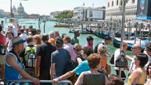Eine Menschenmenge wartet an einem Anleger in Venedig, im Hintergrund sind Gondeln und Gebäude zu sehen.
