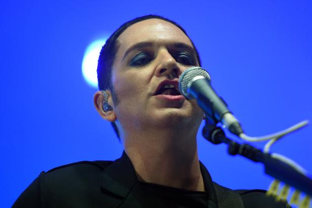 Ein Sänger mit dunklem Make-up singt in ein Mikrofon vor blauem Hintergrund.