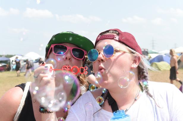 Zwei junge Frauen mit Sonnenbrillen pusten Seifenblasen auf einem Festivalgelände.