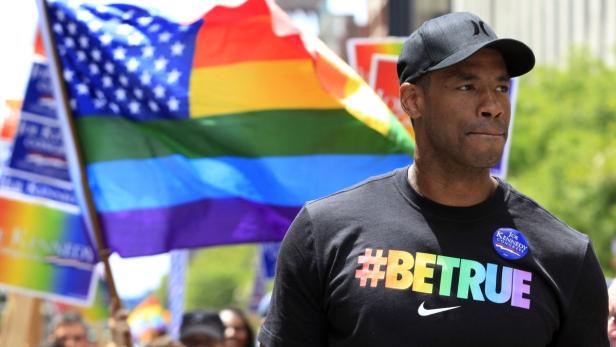 Ein Mann mit Baseballkappe und einem T-Shirt mit der Aufschrift „#BETRUE“ vor einer Regenbogenflagge.