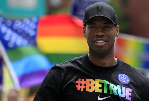 Ein Mann mit einem „#BeTrue“-T-Shirt steht vor einer Regenbogenflagge.