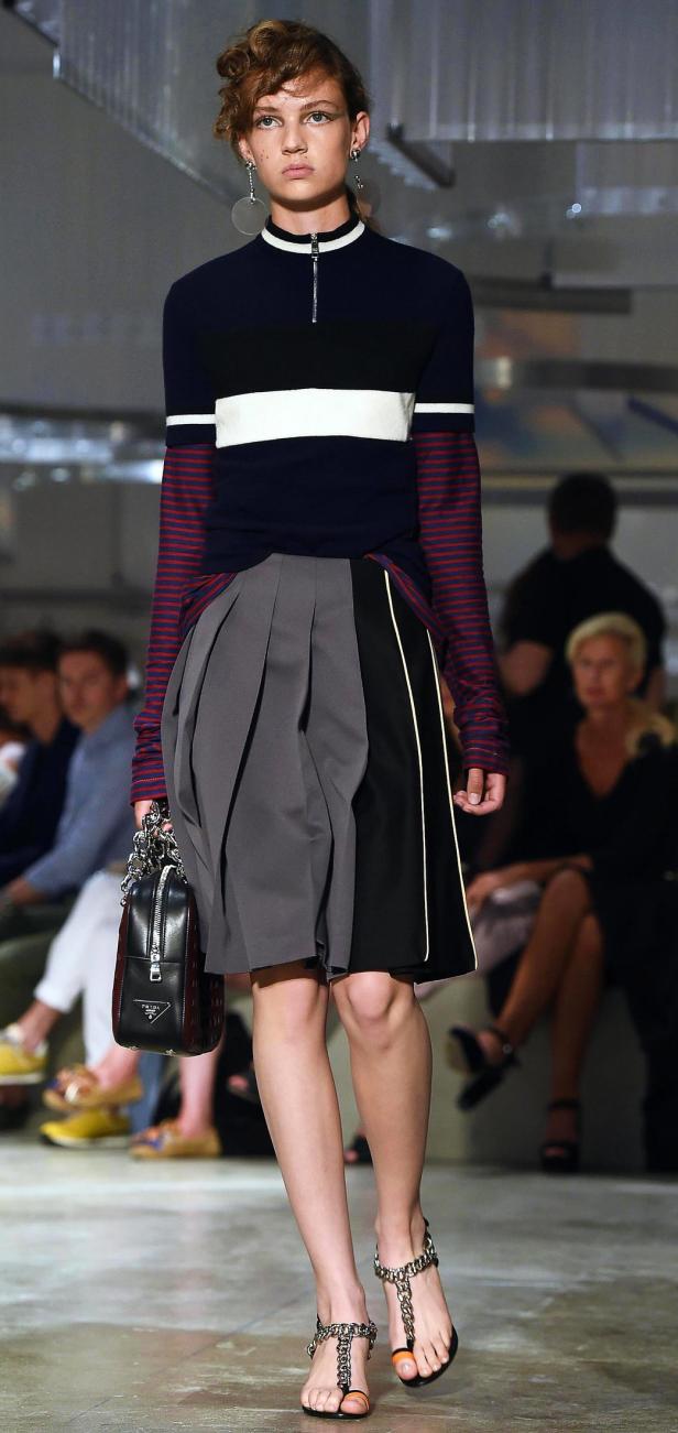 Ein Model präsentiert ein Outfit mit grauem Faltenrock und einer Prada-Handtasche auf dem Laufsteg.