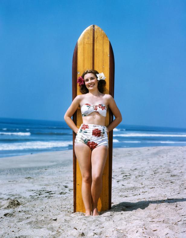 Eine Frau im Bikini mit Blumen im Haar posiert am Strand vor einem Surfbrett.