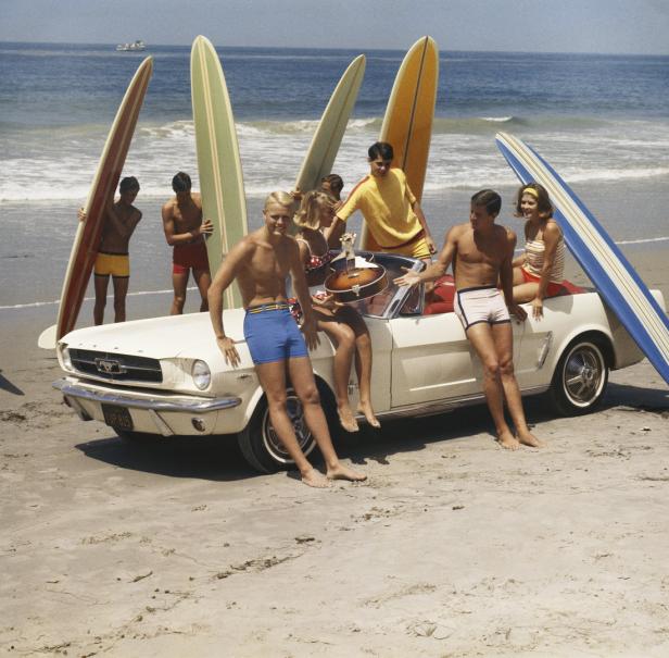 Eine Gruppe junger Leute mit Surfbrettern posiert an einem Strand neben einem weißen Oldtimer-Cabrio.