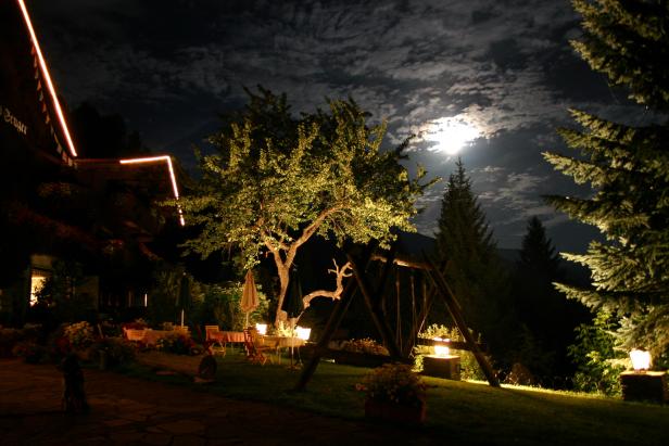 Nachts beleuchteter Garten mit Tischen, Stühlen und einer Schaukel unter einem Baum im Mondschein.