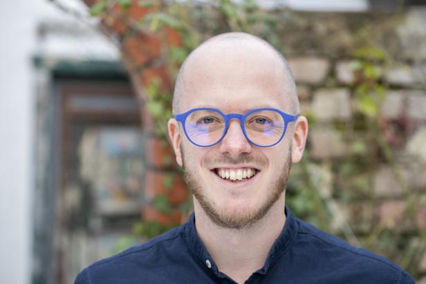 Ein lächelnder Mann mit Bart und blauen Brillengläsern vor einer Ziegelmauer.