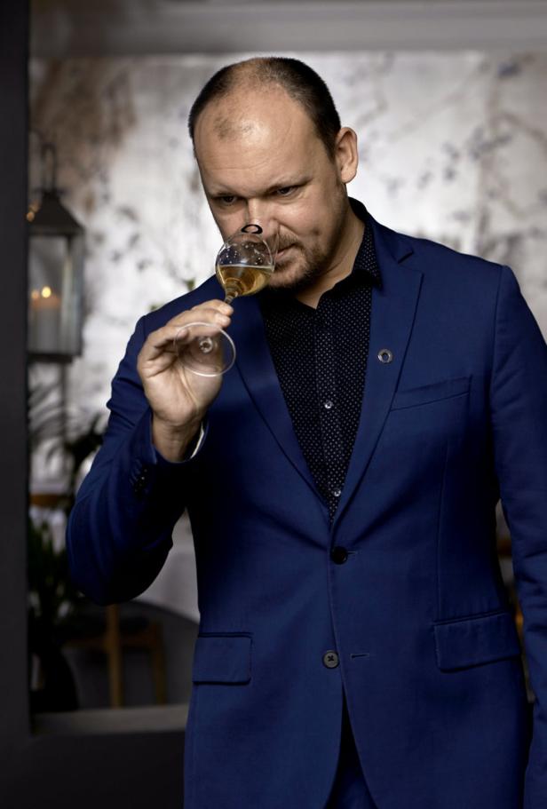 Ein Mann im blauen Anzug riecht an einem Glas mit hellem Getränk.