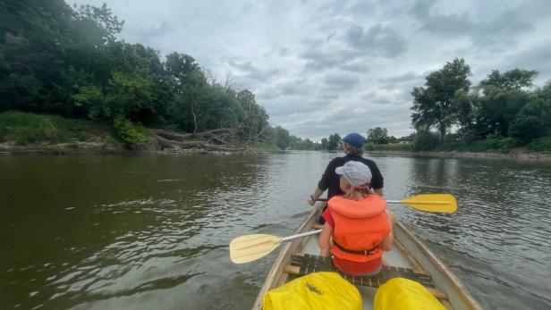Zwei Personen paddeln in einem Kanu auf einem Fluss entlang bewaldeter Ufer.