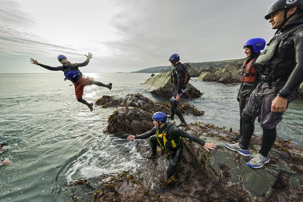 Eine Gruppe von Personen springt von Felsen ins Meer, bekleidet mit Neoprenanzügen und Helmen.