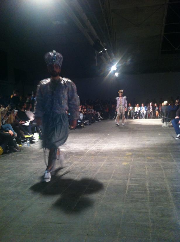 Zwei Models präsentieren extravagante Mode auf einem Laufsteg vor Publikum.