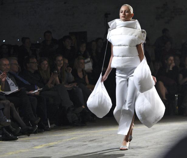 Eine Frau präsentiert auf einem Laufsteg ein weißes, ungewöhnliches Outfit mit großen, sackartigen Accessoires.