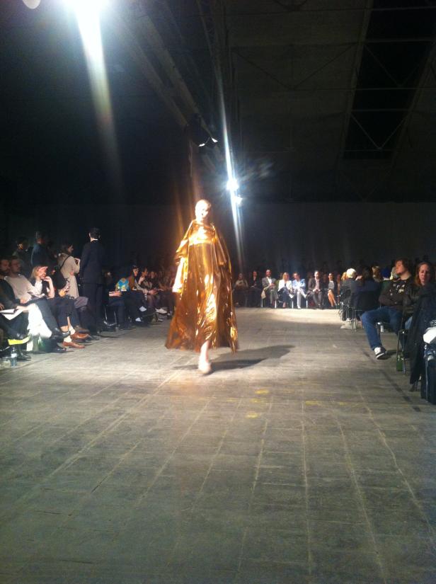 Eine Person in einem goldenen Gewand läuft über einen Laufsteg bei einer Modenschau.