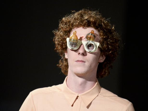 Ein Mann mit lockigem Haar trägt eine extravagante Brille mit Babyflaschenaufsätzen.