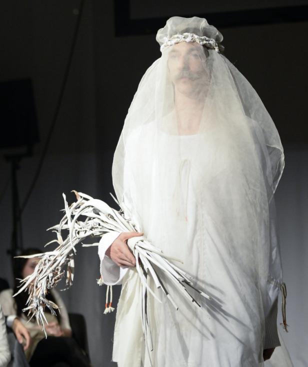 Ein Mann mit Schnurrbart, gekleidet wie eine Braut, hält einen Strauß weißer Zweige.