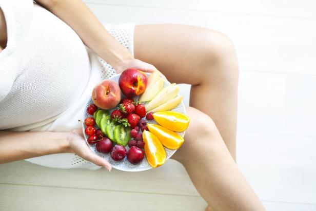 Schwangere Frau mit einem Teller Obst wie Erdbeeren, Kiwis, Pflaumen und Orangen.