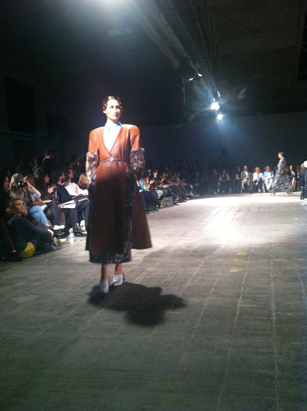 Eine Frau präsentiert ein braunes Kleid mit Fellbesatz auf einem Laufsteg.