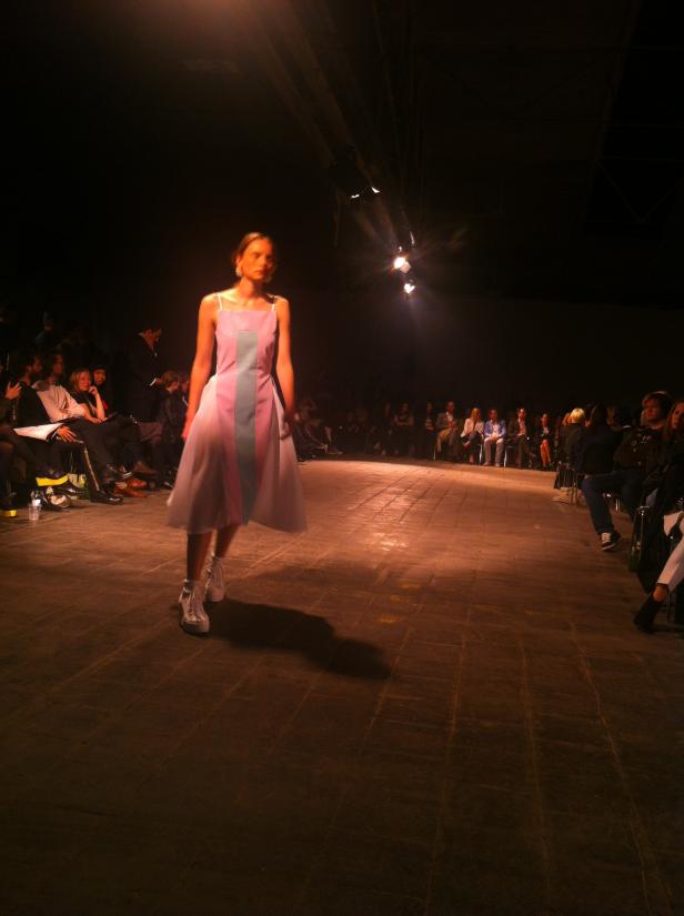 Ein Model präsentiert ein rosa-blaues Kleid auf einem Laufsteg vor Publikum.