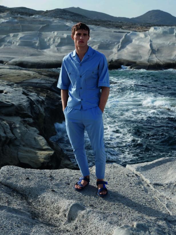 Ein Mann in hellblauer Kleidung und Sandalen steht an einer felsigen Küste.