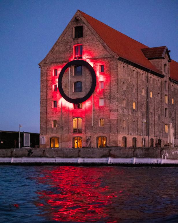 Ein Backsteingebäude mit einem großen schwarzen Kreis, beleuchtet durch rotes Licht, das sich im Wasser spiegelt.