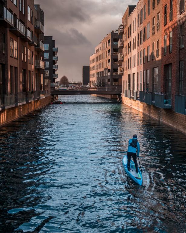 Eine Person paddelt auf einem Stand-Up-Paddleboard durch einen Kanal zwischen Backsteingebäuden.