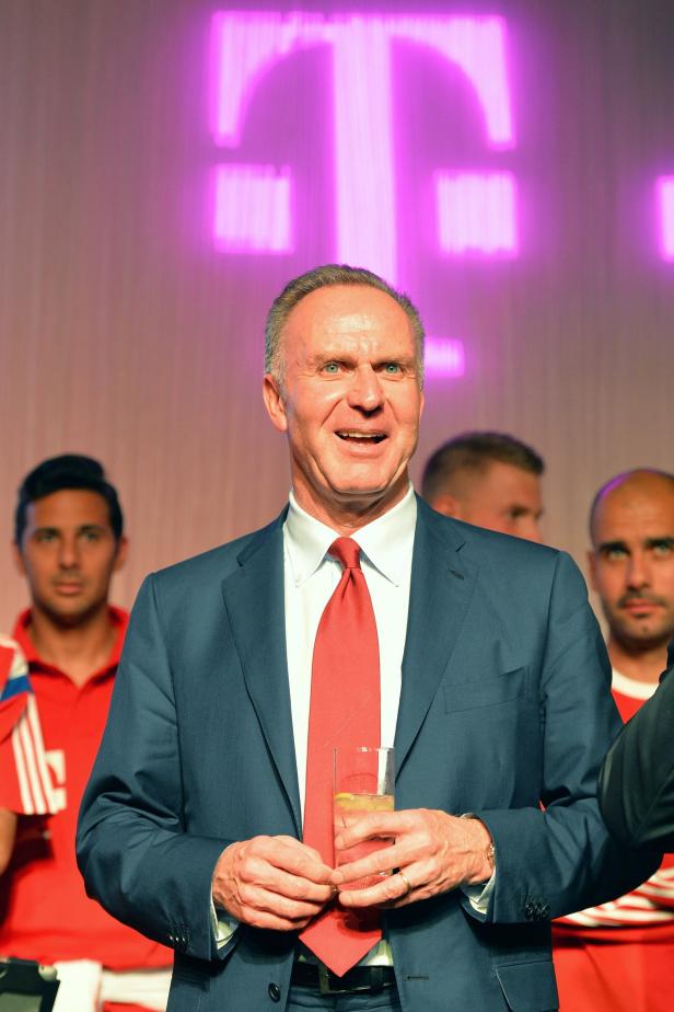 Uli Hoeneß hält ein Glas Wasser vor einem T-Mobile-Logo.