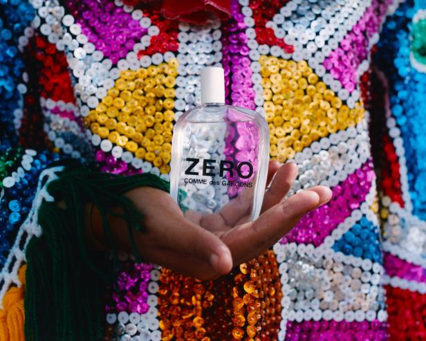 Eine Hand hält eine Flasche „ZERO Comme des Garçons“ vor einem bunten, paillettenbesetzten Hintergrund.