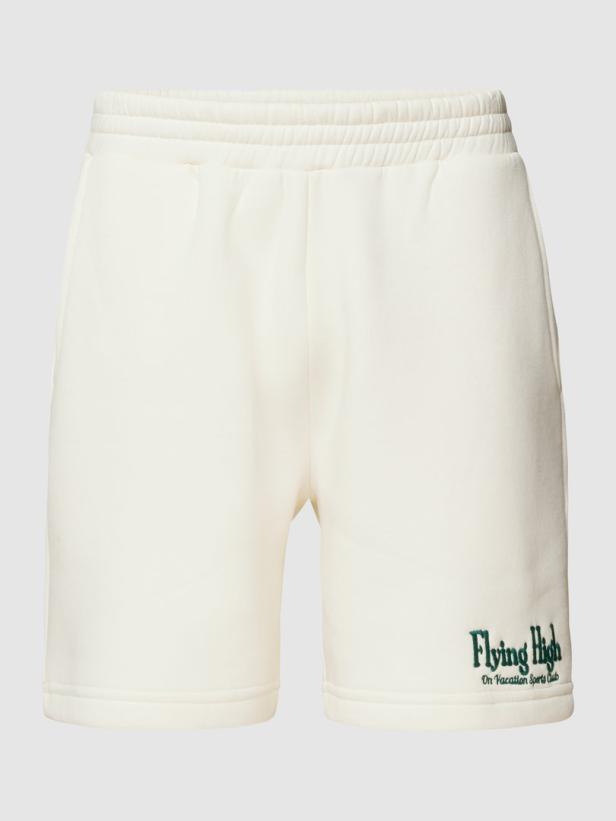 Eine kurze, cremefarbene Hose mit dem grünen Aufdruck „Flying High On Vacation Sports Club“.