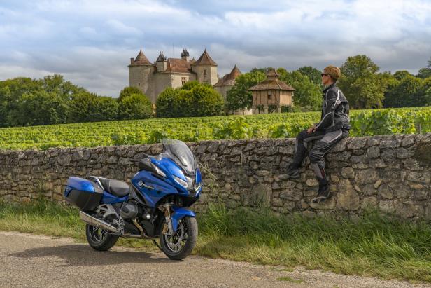 Eine Frau sitzt auf einer Steinmauer neben einem blauen Motorrad vor einem Schloss und einem Weinberg.