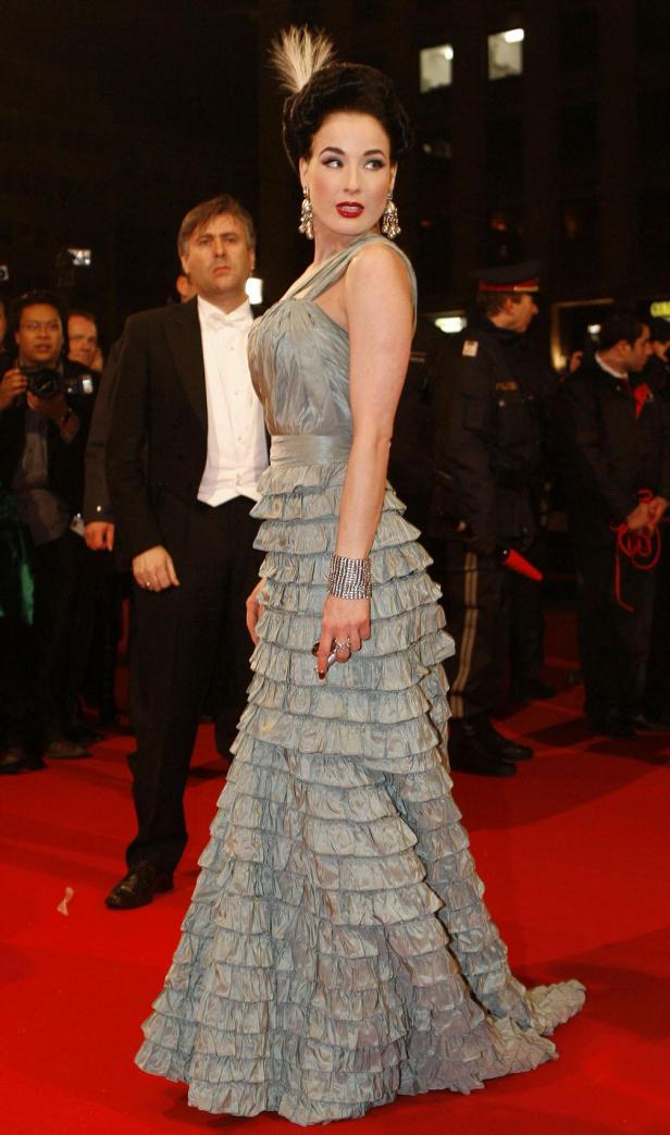 Dita Von Teese in einem grauen Rüschenkleid auf einem roten Teppich.