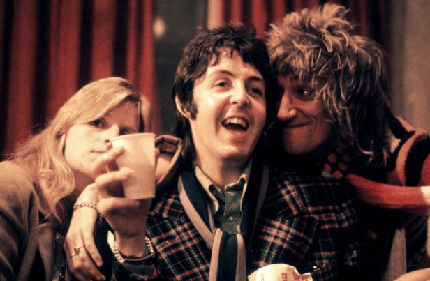 Paul McCartney umarmt mit Rod Stewart und einer Frau mit einem Becher in der Hand.