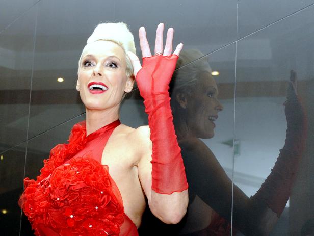 Brigitte Nielsen in einem roten Kleid mit passenden Handschuhen winkt.