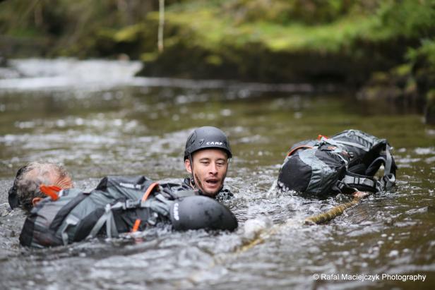 Zwei Personen mit Rucksäcken waten durch einen Fluss.