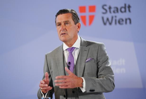 Ein Mann in einem grauen Anzug gestikuliert vor dem Logo der Stadt Wien.