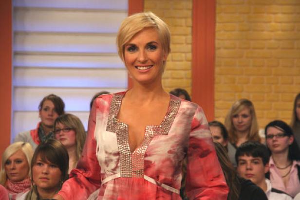 Eine blonde Frau in einem rot-weißen Kleid steht vor einem Publikum.