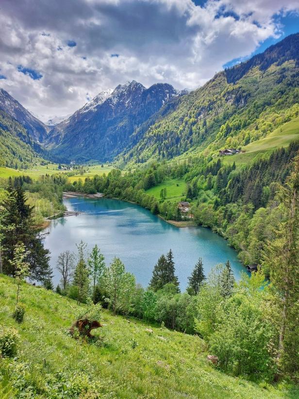 Ein Bergsee liegt eingebettet in einer grünen Hügellandschaft mit schneebedeckten Gipfeln im Hintergrund.
