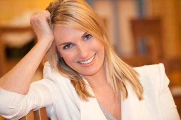Eine lächelnde Frau mit blonden Haaren und weißem Blazer posiert für ein Porträt.