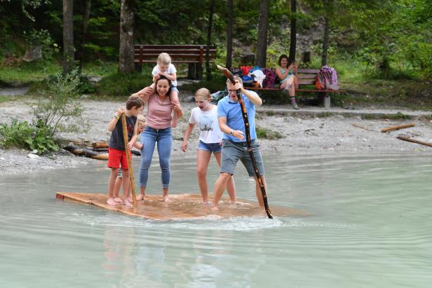 Eine Familie steht auf einem Floß in einem See und stakt mit Stöcken.