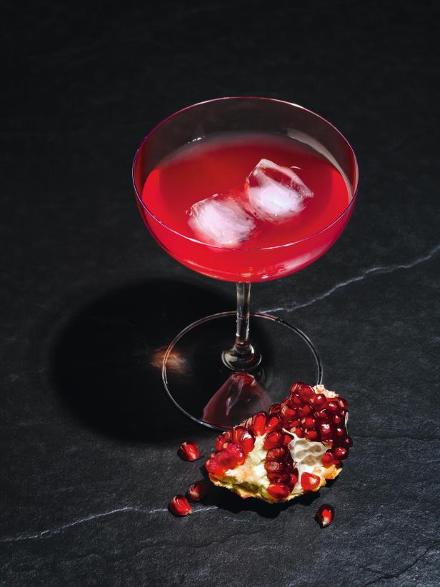 Ein roter Cocktail mit Eiswürfeln und einer Granatapfelspalte auf einem dunklen Tisch.